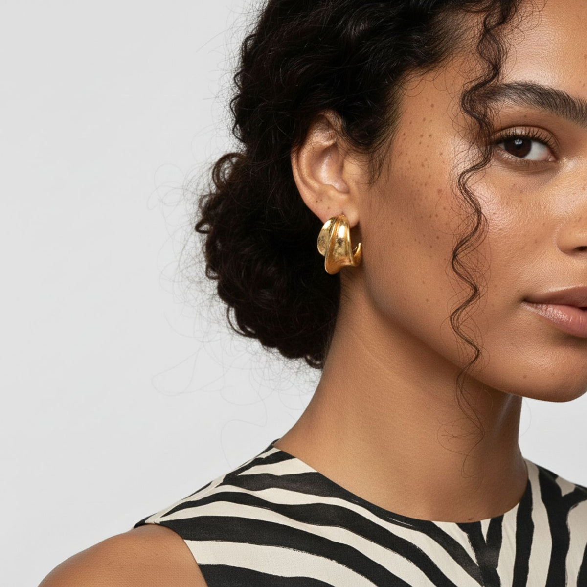 A woman with curly dark hair wears a zebra-print sleeveless top and JAREDJAMIN Jewelry Online’s Cressida Gold Hoop Earrings. The image highlights her glowing skin, subtle makeup, and a wavy strand of hair on her cheek.