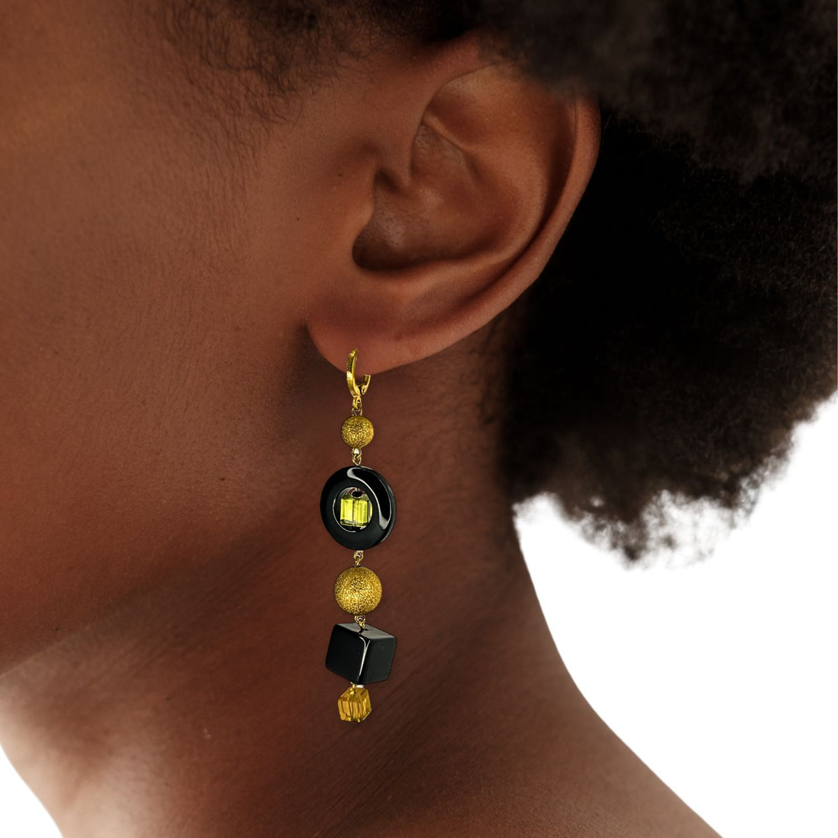Un person con short, curly hair y brown skin wears JAREDJAMIN Joyería Online's Cubism Leverback colgante pendientes, con geometric glass beads, oro y black accents, faceted yellow bead, y 18K bañado en oro leverbacks.