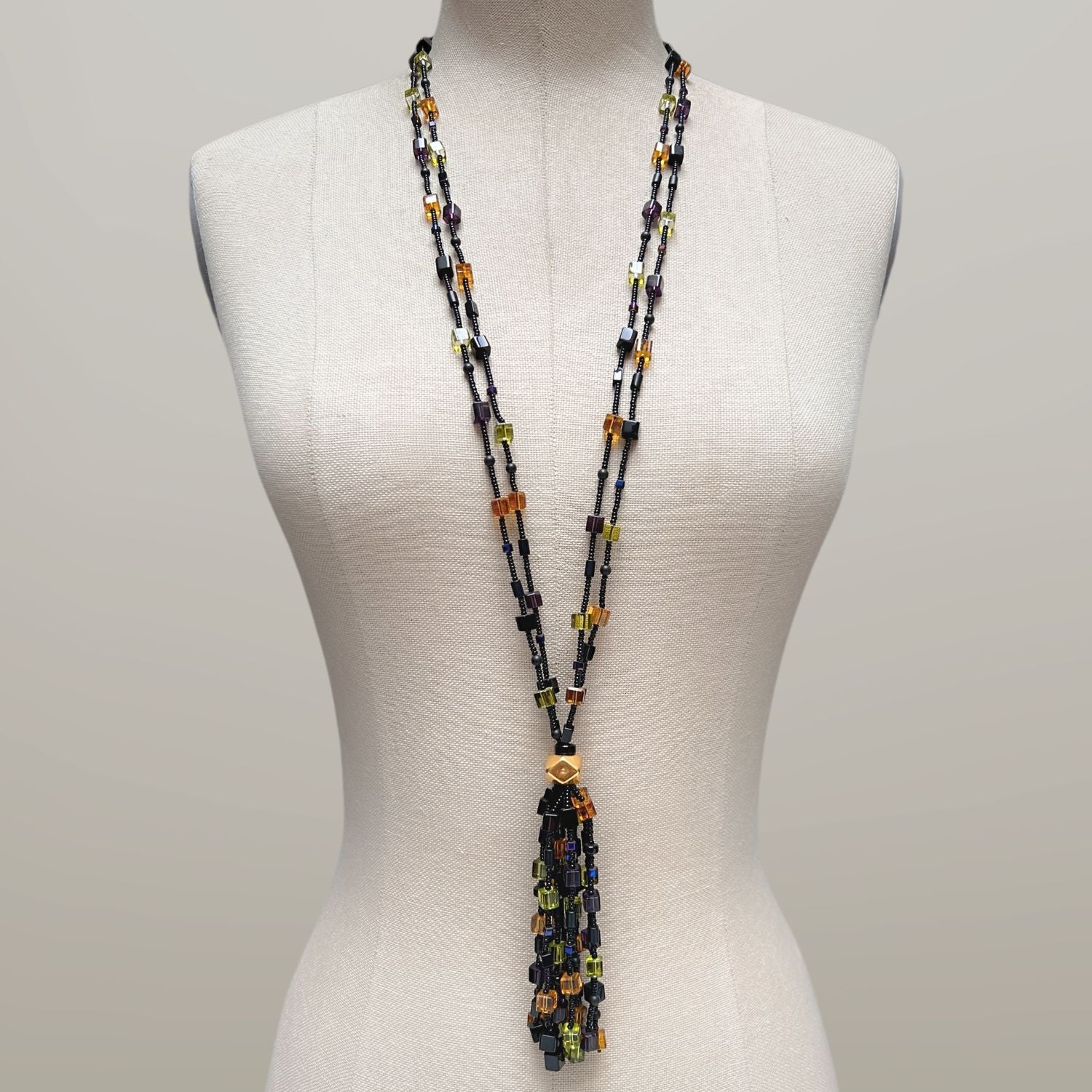 The Cubism Tassel Necklace by JAREDJAMIN Jewelry Online features two black strands with yellow, orange, green, and purple beads, joined at a geometric gold bead and finished with a matching tassel and gold toggle clasp.