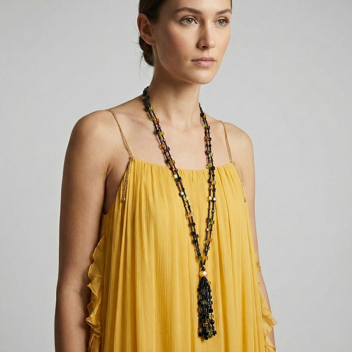 Un woman dans un sleeveless yellow dress stands against a plain backdrop, wearing the Cubism Tassel collier par JAREDJAMIN Bijoux en Ligne—its long tassel et geometric or beads accent her look as she gazes calmly ahead.