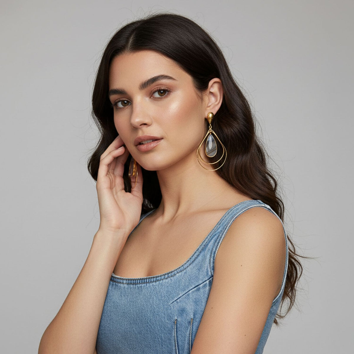 Un woman avec long, wavy dark hair models the Darlena or Agate pendante boucles d'oreilles par JAREDJAMIN Bijoux en Ligne. Wearing a sleeveless denim top, she touches her face et looks at the camera, her natural makeup glowing against a light gray background.