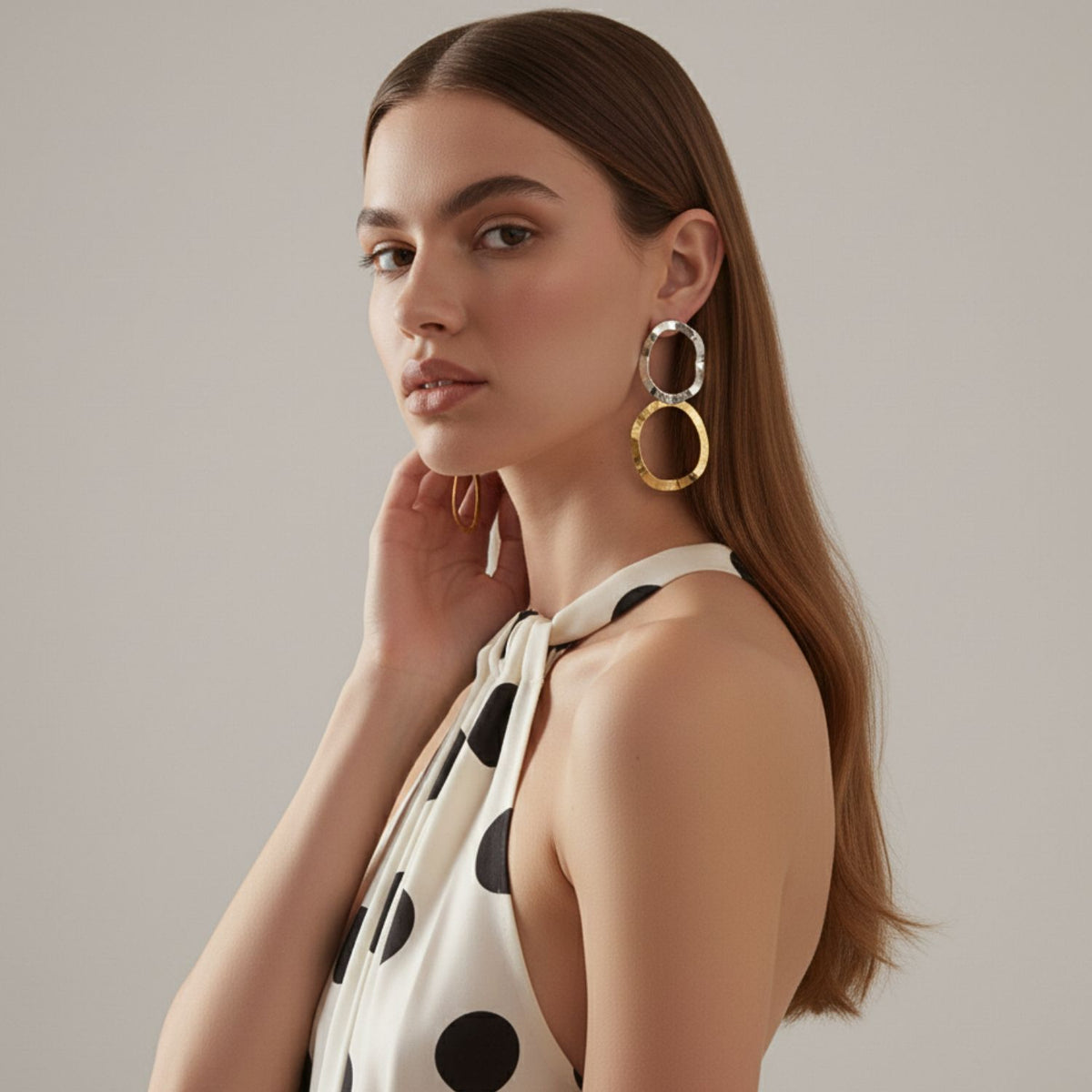 Un woman con long, straight brown hair in profile wears a sleeveless white dress con black polka dots y JAREDJAMIN Joyería Online's Duet colgante Post pendientes—audaz, tarnish-free 18K oro double circles—her hand gently touching her neck.