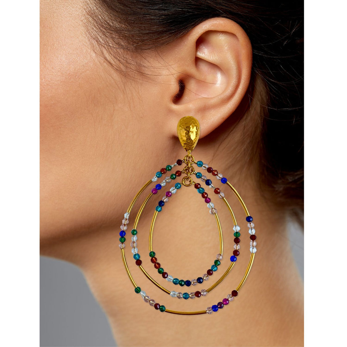 A close-up features Easy Breezy Multicolor Post Earrings by JAREDJAMIN Jewelry Online, with large oval-shaped tiers crafted in 18K gold plate. The earrings display vibrant blue, green, red, and clear bead embellishments and a hammered gold texture on top against a gray background.