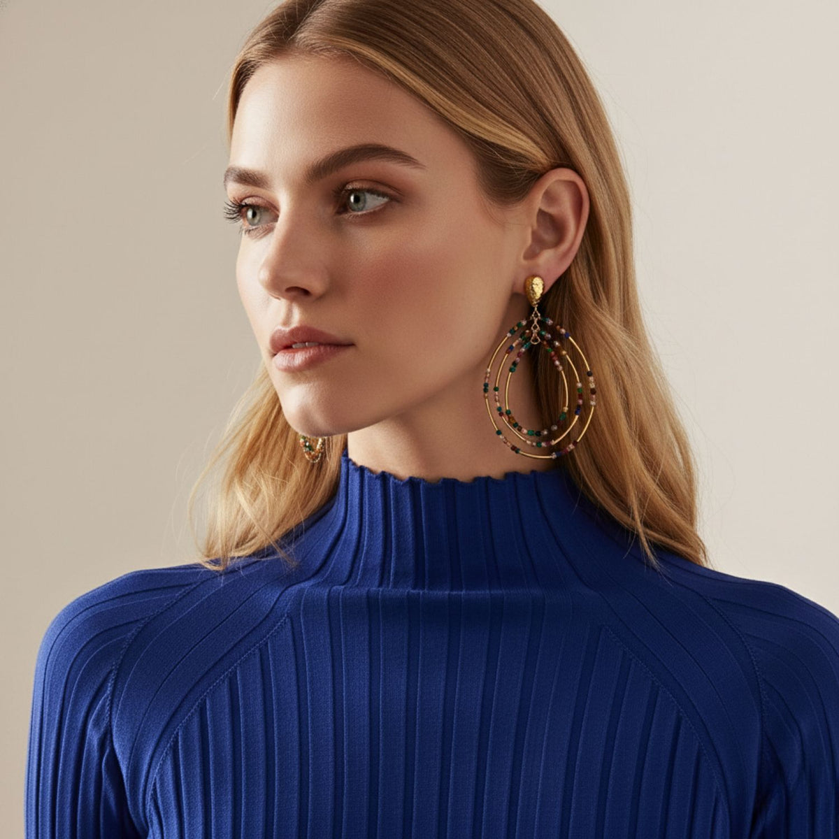 A woman with long blonde hair wears a royal blue, ribbed high-neck top and JAREDJAMIN Jewelry Online's Easy Breezy Multicolor Post Earrings, which add subtle sparkle as she gazes calmly to the side against a neutral background.