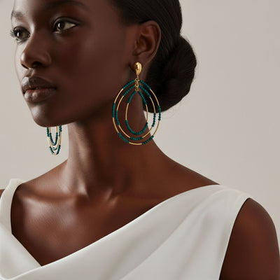 A woman with dark skin and sleek hair wears a white sleeveless top, her look accented by JAREDJAMIN Jewelry Online's Easy Breezy Turquoise Post Earrings. The neutral background enhances her elegant style and the vivid teal beads of the statement earrings.