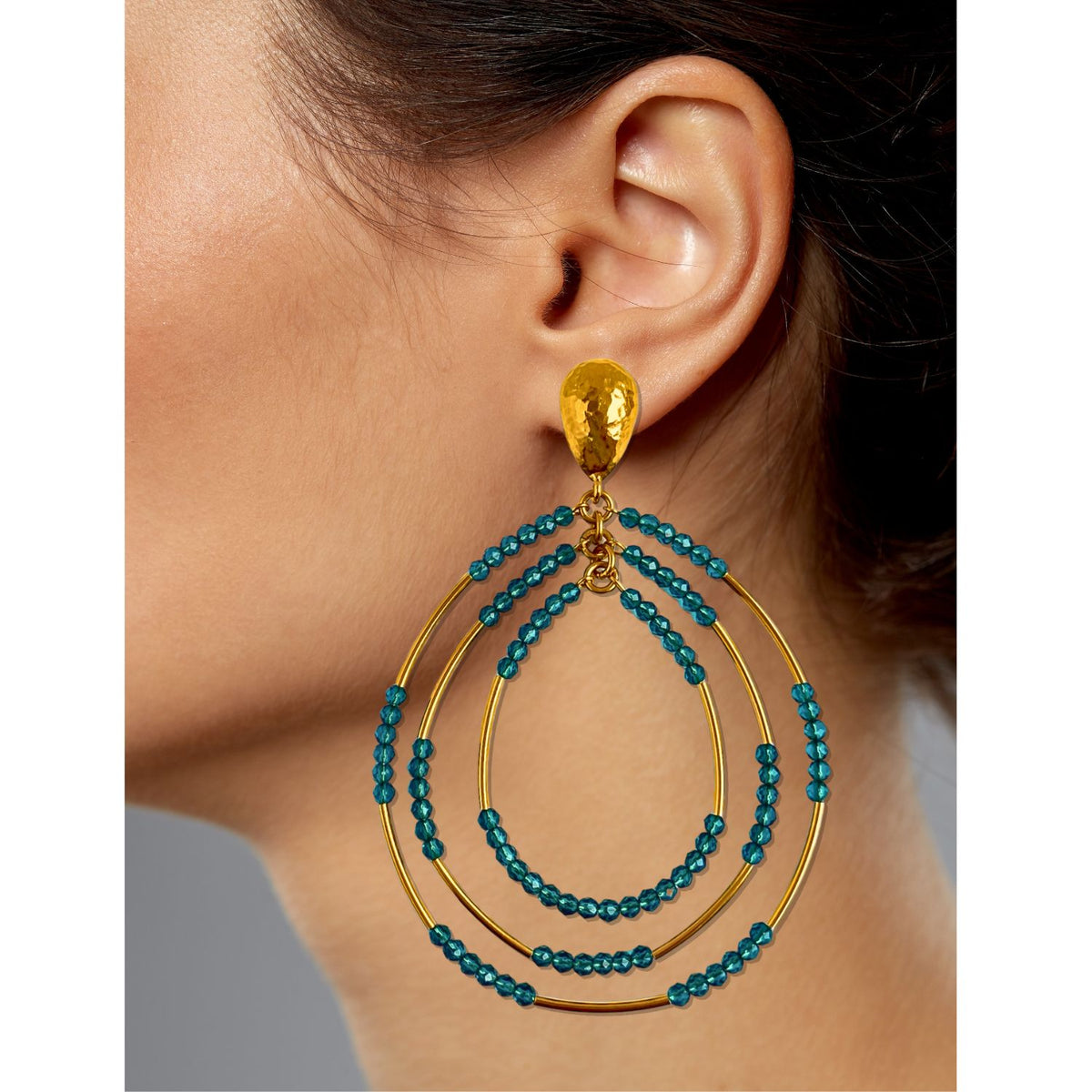 Close-up of a woman&#39;s ear wearing Easy Breezy Turquoise Post Earrings from JAREDJAMIN Jewelry Online. The earrings feature gold hoops with small turquoise crystals, attached by a textured gold stud. A softly blurred background emphasizes the intricate design.