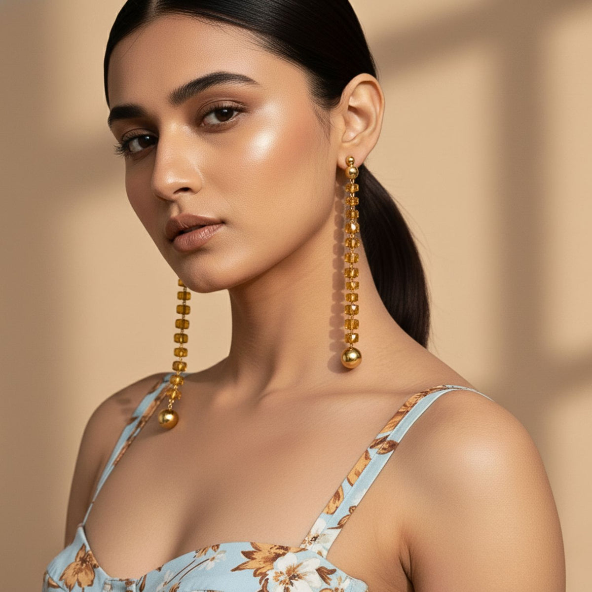 Un woman avec smooth, glowing skin et straight dark hair wears striking Erica Dangle Post boucles d'oreilles par JAREDJAMIN Bijoux en Ligne, paired avec a blue floral spaghetti-strap top. She gazes confidently at the camera against a softly-lit beige background.