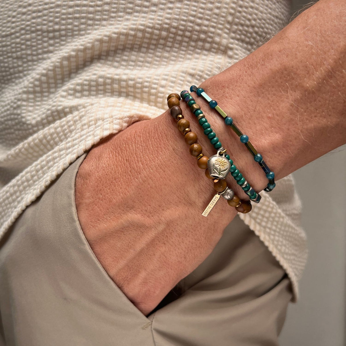 A person in beige pants and a textured cream shirt displays the Everest Foothill Bracelet Set by JAREDJAMIN Jewelry Online, featuring brown, teal, and silver gemstone beads plus a charm with a rectangular metal tag.