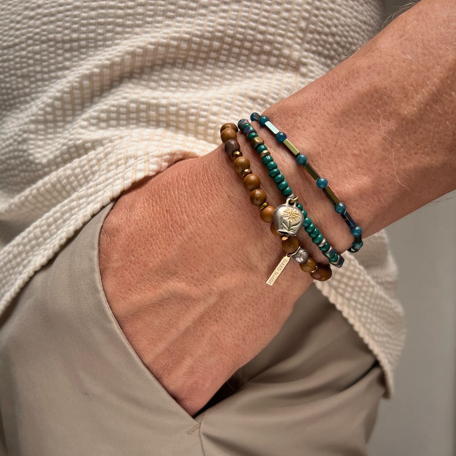 The Everest Foothill Bracelet Set by JAREDJAMIN Jewelry Online features coiled Verdigris beads with green, brown, and turquoise accents, plus a silver lotus charm. Displayed on a plain white background.