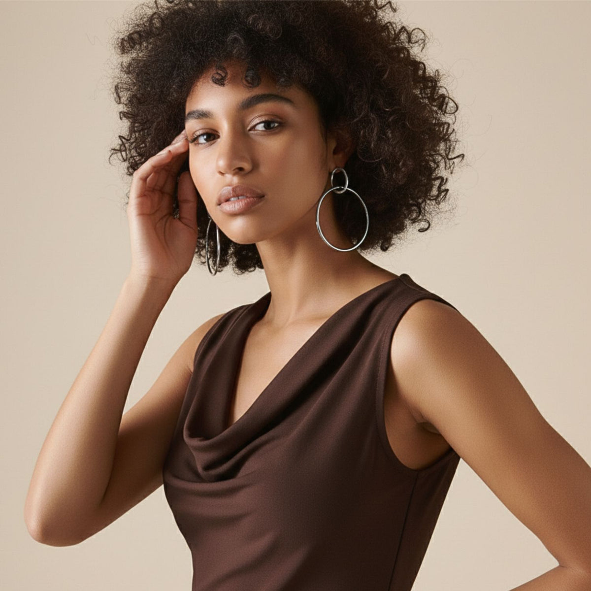 A young woman confidently poses against a beige background, wearing JAREDJAMIN Jewelry Online&#39;s Everly Dangle Hoop Post Earrings and a sleeveless brown draped-neck top, her hand touching her curly hair for an elegant portrait.