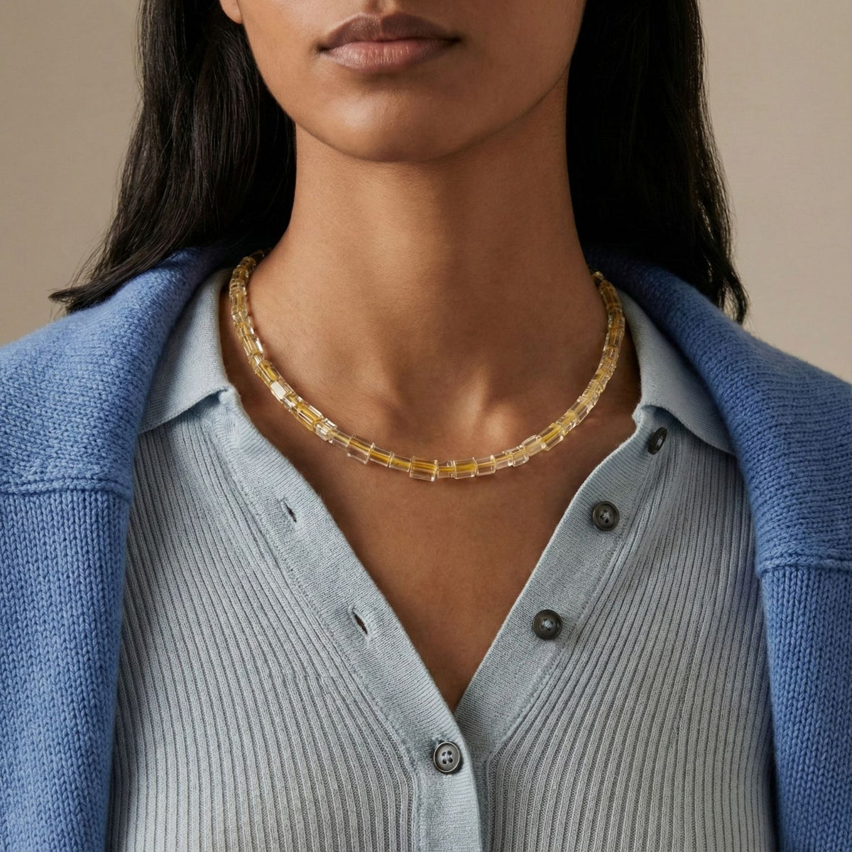 A woman in a light blue ribbed shirt and blue sweater wears JAREDJAMIN Jewelry Online’s Faceted Citrine Bead Necklace, featuring chunky translucent beads. She has straight dark hair and is posed against a neutral background.