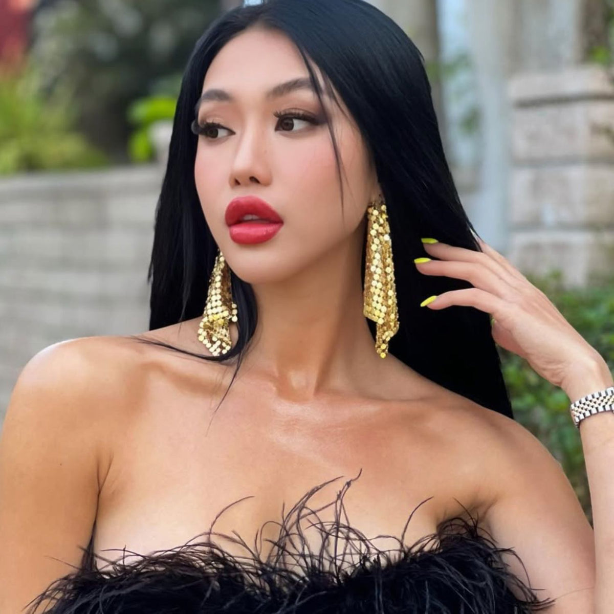 A person with long black hair and striking makeup poses outdoors, exuding elegance while wearing Fazzoletto Chainmail Post Earrings from JAREDJAMIN Jewelry Online and a top adorned with black feathers, complemented by a metallic mesh watch. A blurred backdrop of greenery and stone structures highlights their confident expression.