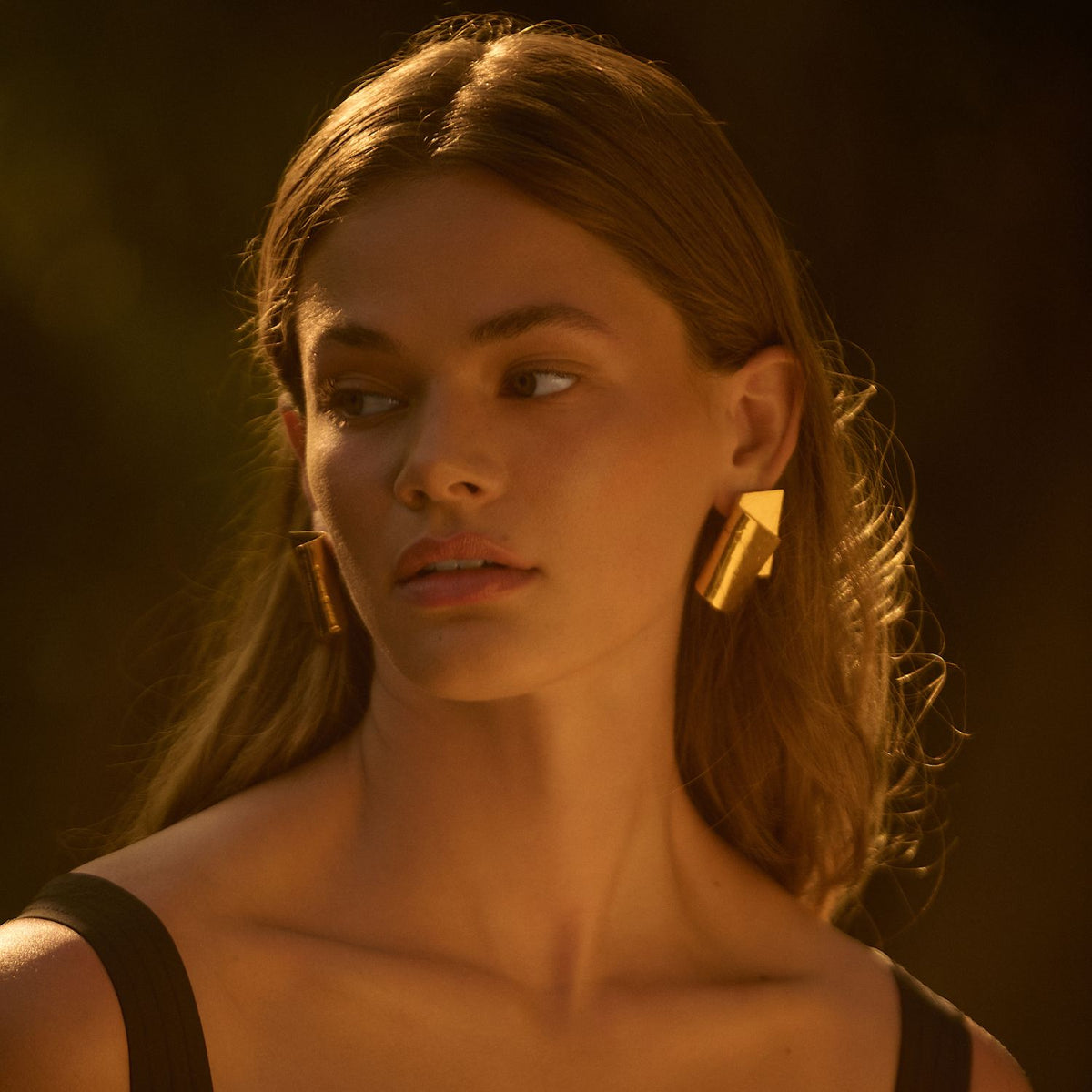 Un young woman avec wavy brown hair gazes to the side in soft light, wearing a black top et or Fold Over Post boucles d'oreilles de JAREDJAMIN Bijoux en Ligne. Les dark, blurred background highlights her calm look et the boucles d'oreilles’ radiant or shine.
