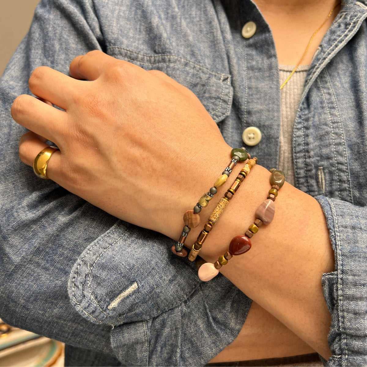 A person の中の ブルー denim shirt crosses their arms, showcasing Forest Floor ブレスレット Set JAREDJAMIN製 ジュエリーオンライン—three beaded ブレスレットとjasperとmulticolored stones, plus a ゴールド リングとa thin ゴールド チェーン around their neck.