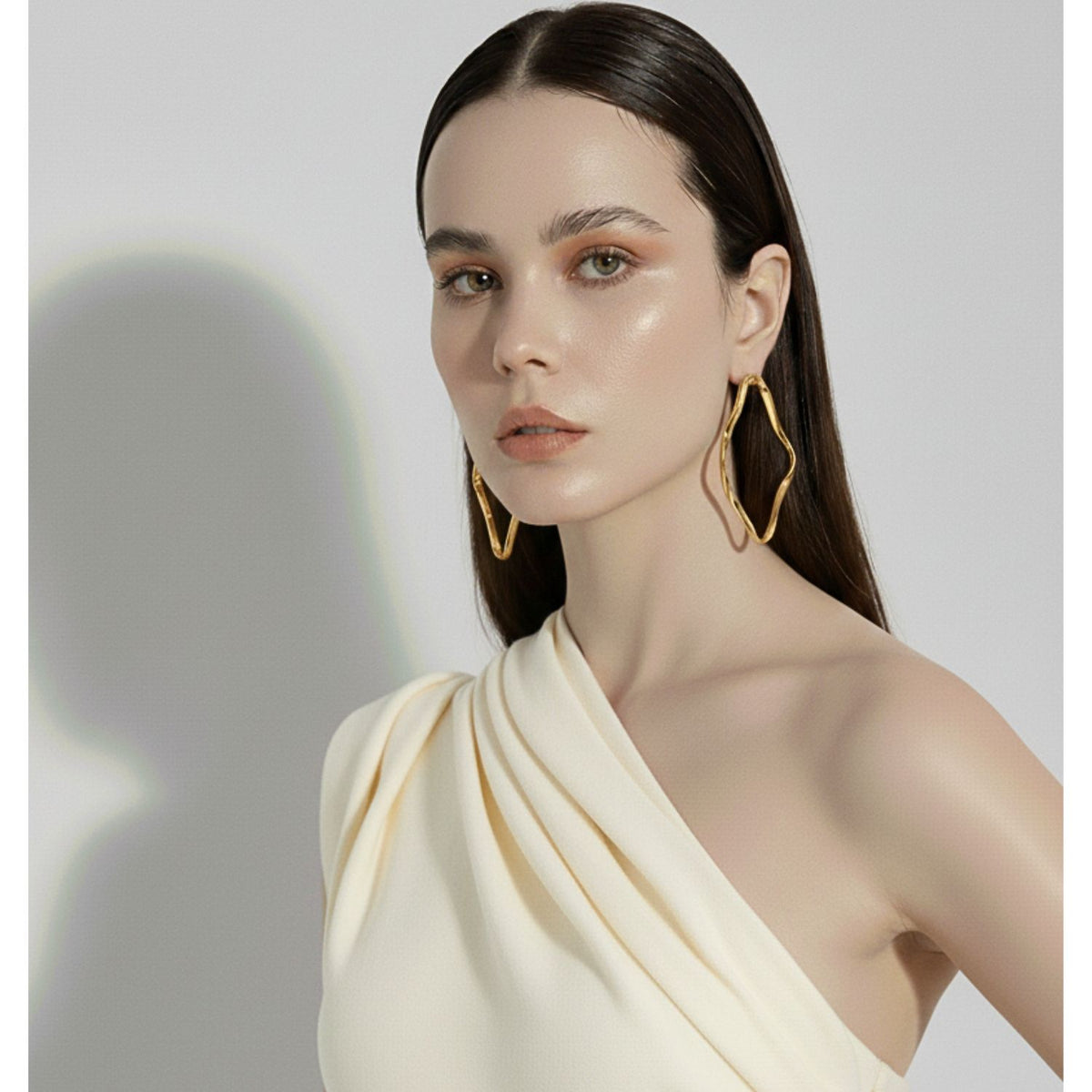 A woman with straight dark brown hair wears a cream one-shoulder top and JAREDJAMIN Jewelry Online’s Gold Ribbonelle Post Earrings. Her makeup is natural and dewy, and she faces forward against a plain light background with a soft shadow.
