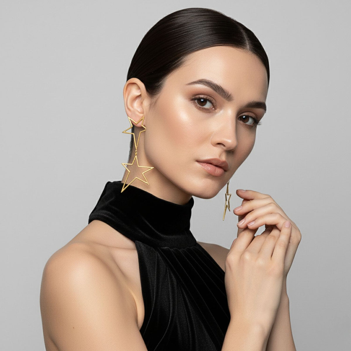 Un woman avec smooth dark hair dans un low bun wears a black high-neck sleeveless top, posing avec her hands near her face to showcase JAREDJAMIN Bijoux en Ligne’s or Shooting Star Dust Dangle Post boucles d'oreilles against a light gray background.