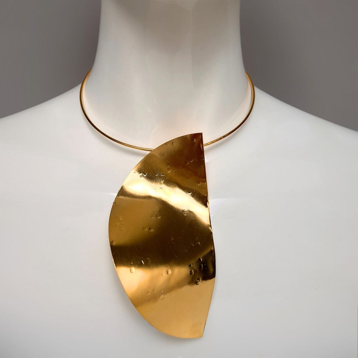 The Geo Twist Pendant Neck Wire by JAREDJAMIN Jewelry Online is displayed on a mannequin, showcasing a wide, hammered brass half-circle pendant with intricate texture. It hangs from an 18K gold-plated choker for a modern look against a plain gray backdrop.