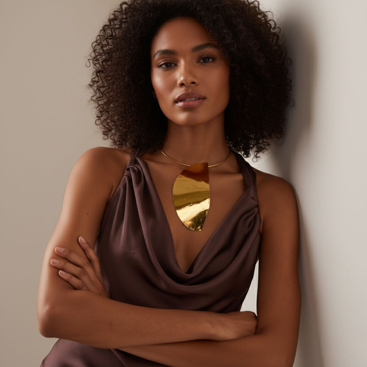 A woman with curly hair, arms crossed, leans on a light wall. She wears a brown sleeveless top and the Geo Twist Pendant Neck Wire by JAREDJAMIN Jewelry Online—its shiny gold design pops against the neutral background.