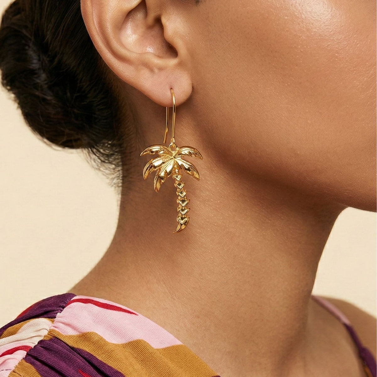 A close-up shows a woman wearing shiny Gold Angeleno Palm Hook Earrings by JAREDJAMIN Jewelry Online. Her hair is tied back, she wears a colorful pink, purple, and yellow patterned top, set against a plain, light beige background.