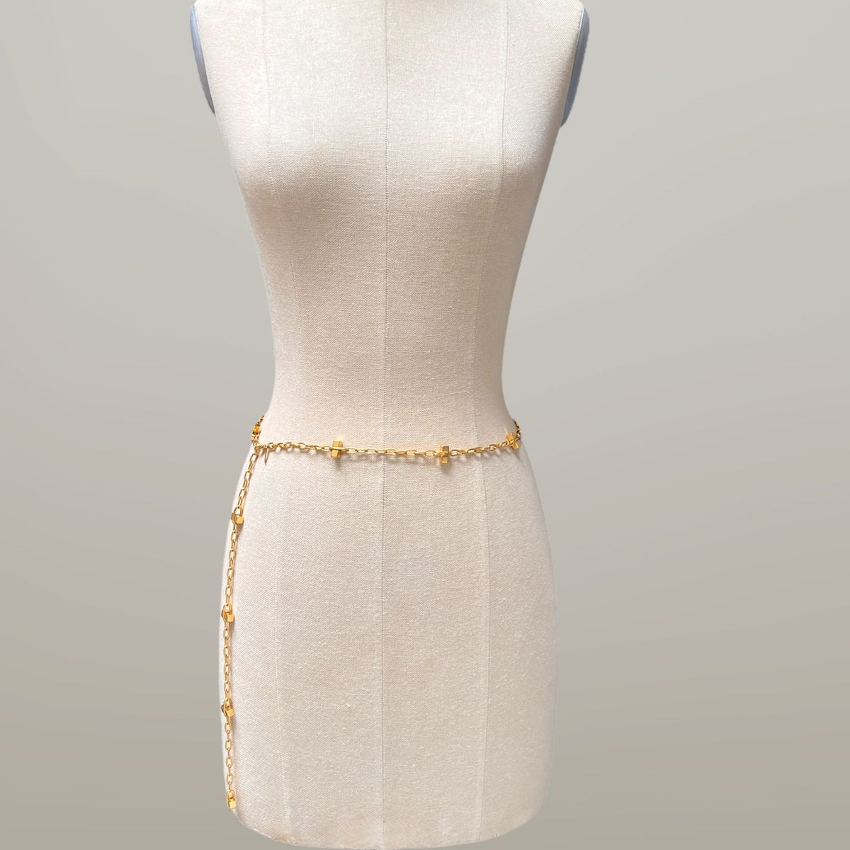 Un dress form mannequin displays the oro Blockbuster Lite Long collar de JAREDJAMIN Joyería Online—a thin, 18K oro plated cadena con evenly spaced oro dijes, draped loosely at the waist against a plain light beige background.