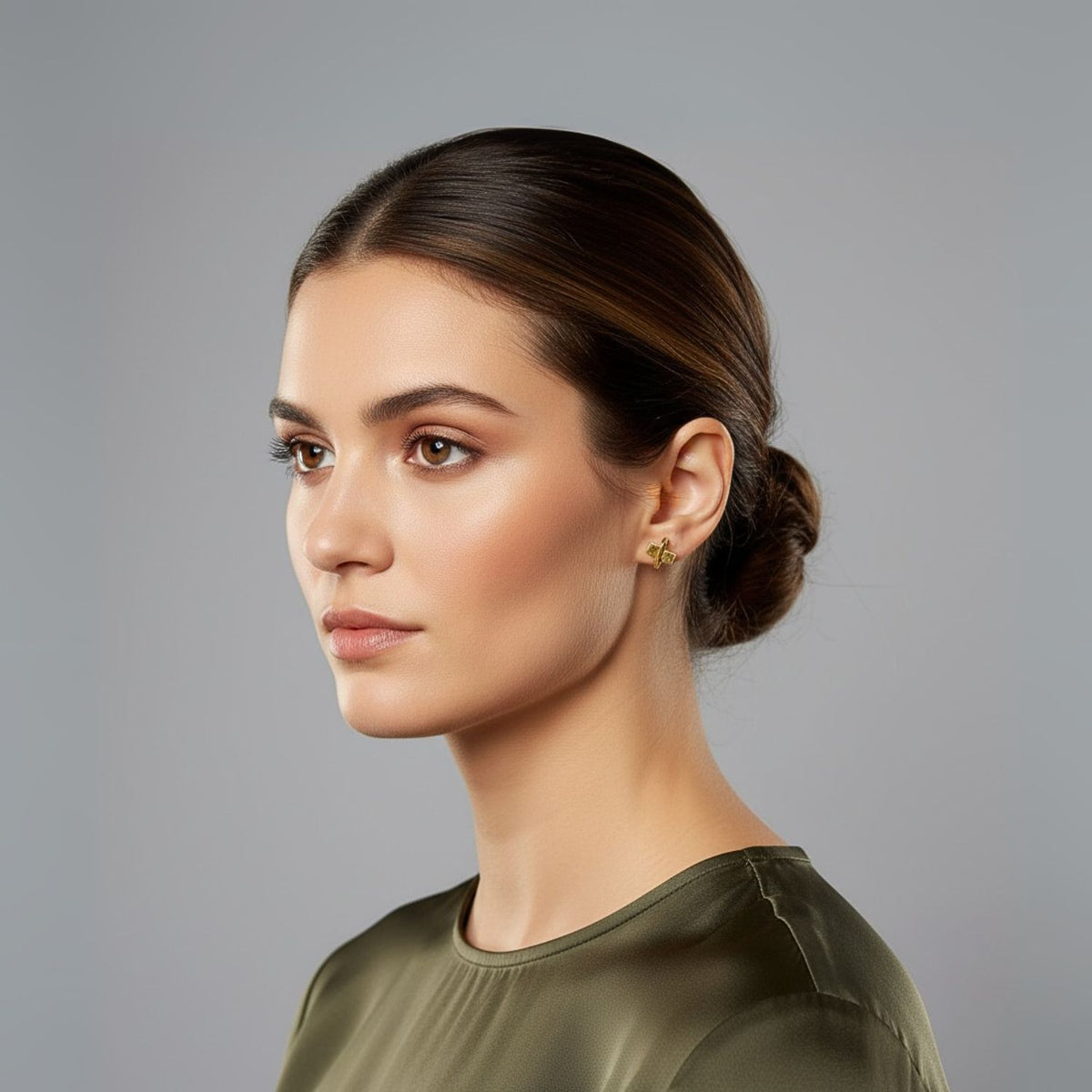 Un woman avec smooth, medium skin tone et dark brown hair dans un sleek low bun looks left. She wears subtle makeup, or Blockbuster puce Post boucles d'oreilles par JAREDJAMIN Bijoux en Ligne, et an olive green satin top before a light gray gradient backdrop.