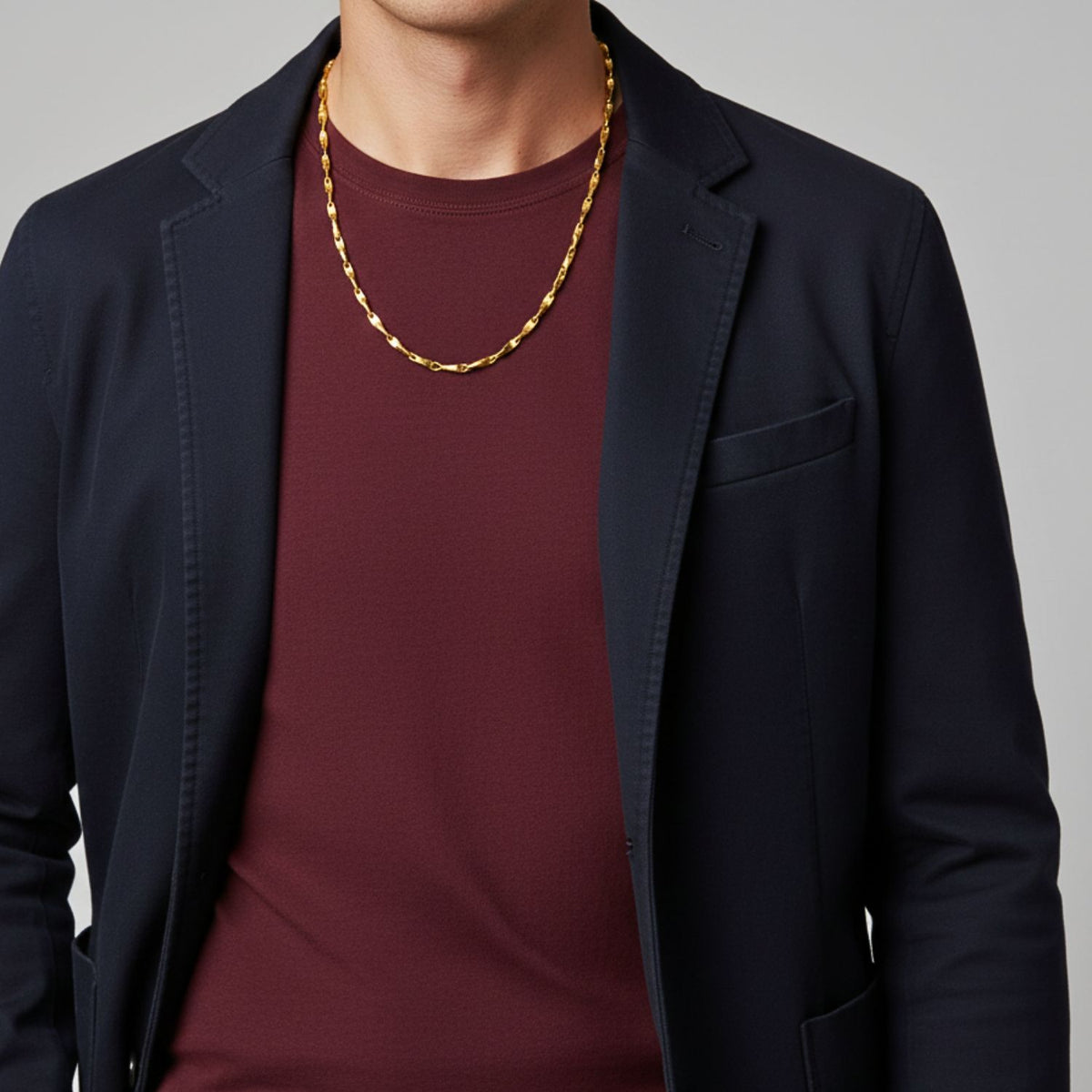 A person wearing a dark blazer over a burgundy shirt showcases the Gold Bobby Bar-Link Chain Necklace by JAREDJAMIN Jewelry Online. The image, cropped at the neck and waist, focuses on the upper torso against a neutral gray background.