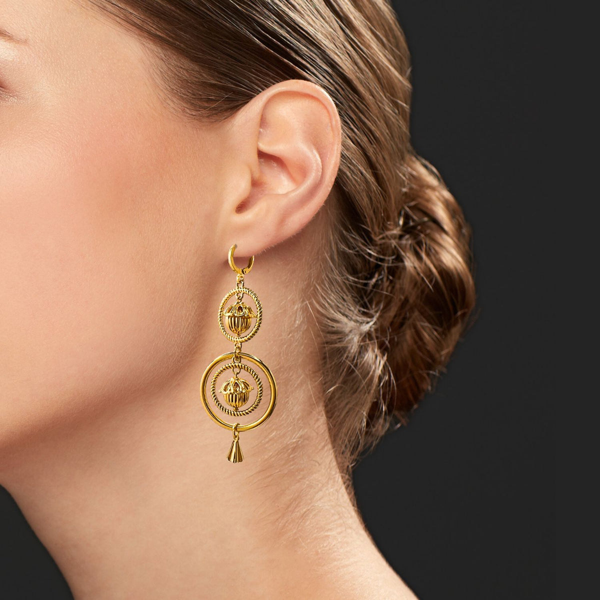 A woman with light skin and brown hair in a low bun wears JAREDJAMIN Jewelry Online’s Gold Bollywood Dangle Leverback Earrings against a dark grey background, highlighting her ear, jawline, and the intricate earring design.
