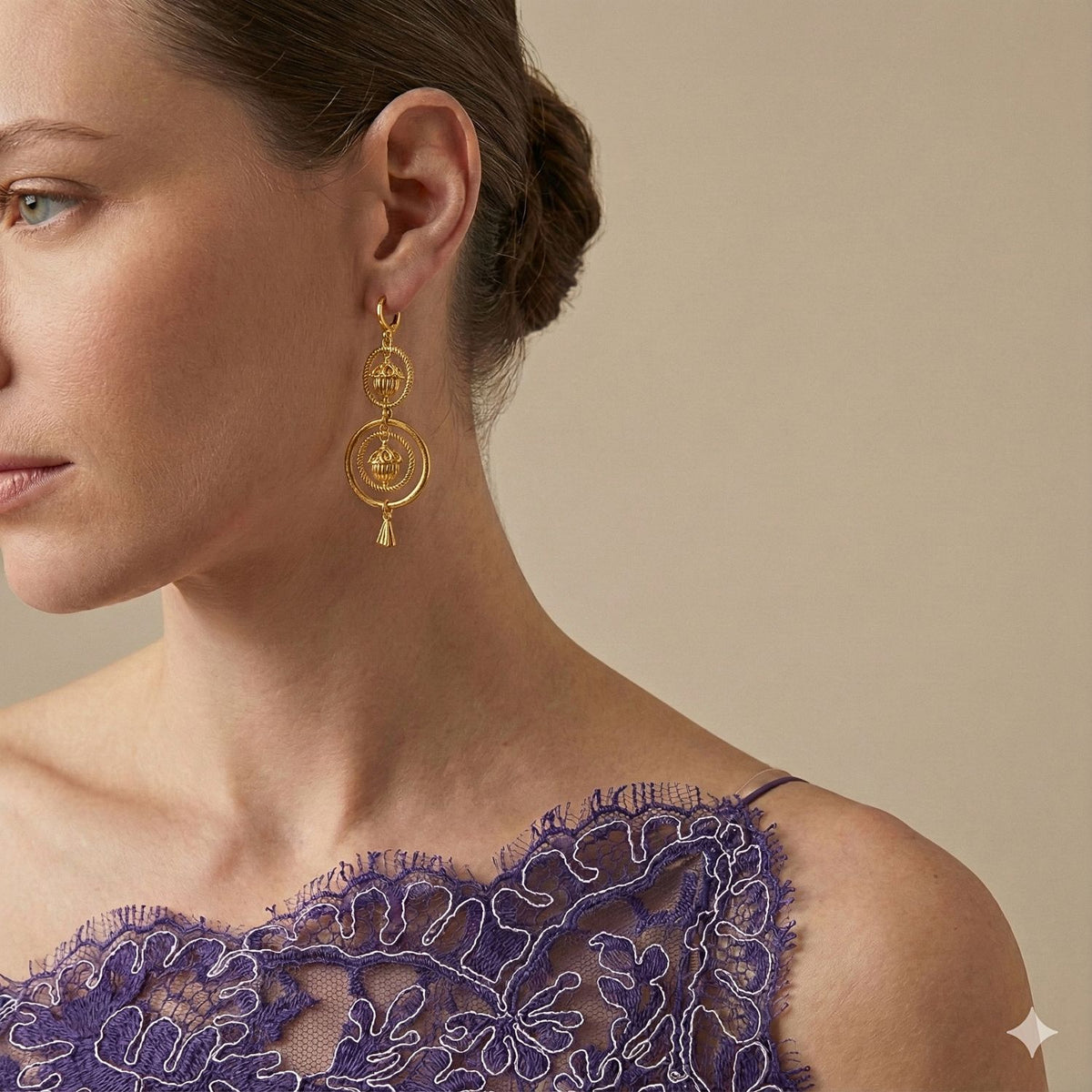 A woman with fair skin and light brown hair in a ponytail wears a sleeveless purple lace dress and JAREDJAMIN Jewelry Online’s Gold Bollywood Dangle Leverback Earrings, featuring circular and tassel details. The background is beige.