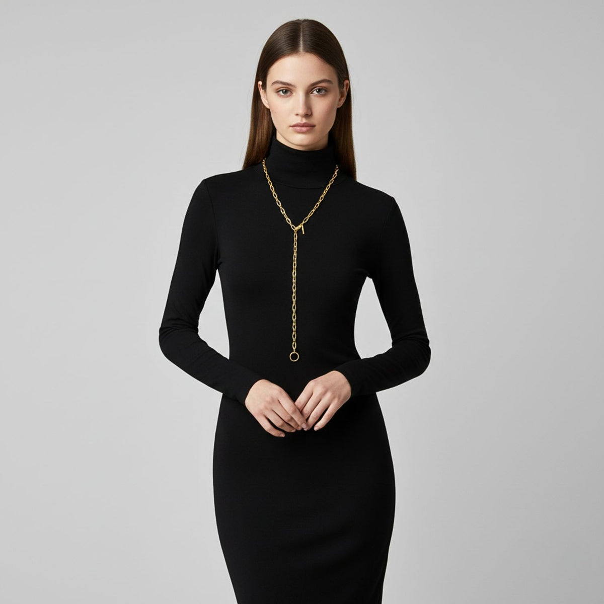 A woman with straight brown hair wears a fitted, long-sleeved black turtleneck dress and the JAREDJAMIN Jewelry Online Gold Chain Reaction Necklace. She stands facing forward against a light gray background, hands clasped at her waist.