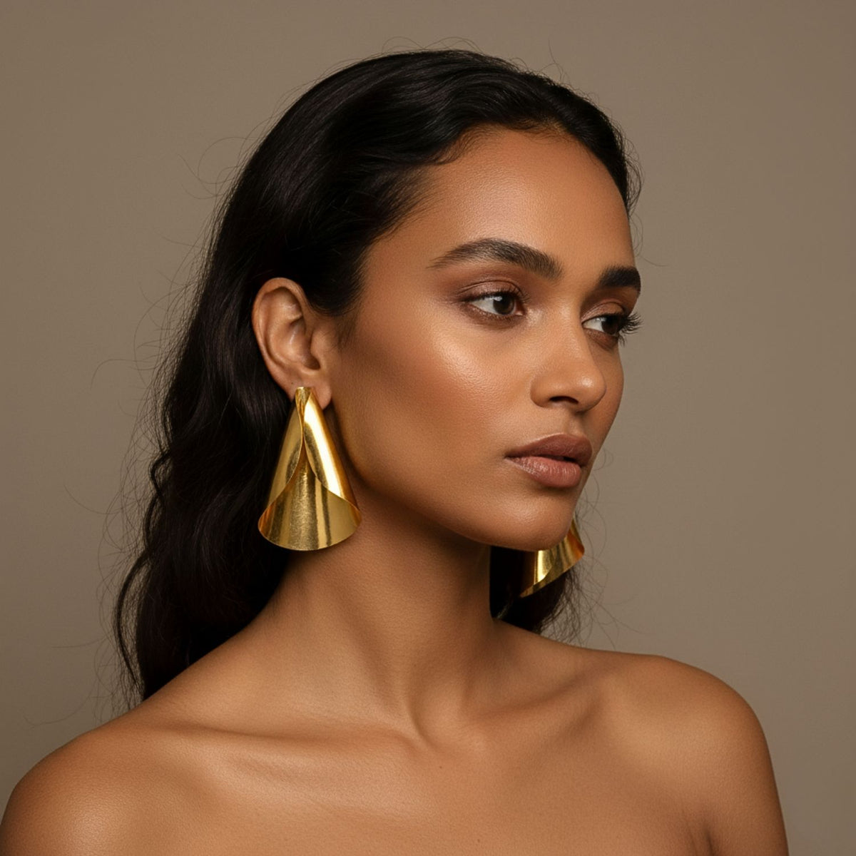 A woman with long, wavy dark hair gazes to the side against a taupe background, wearing JAREDJAMIN Jewelry Online's Gold Connie Post Earrings—striking pieces crafted from 18K gold-plated hammered brass—highlighting her natural elegance.