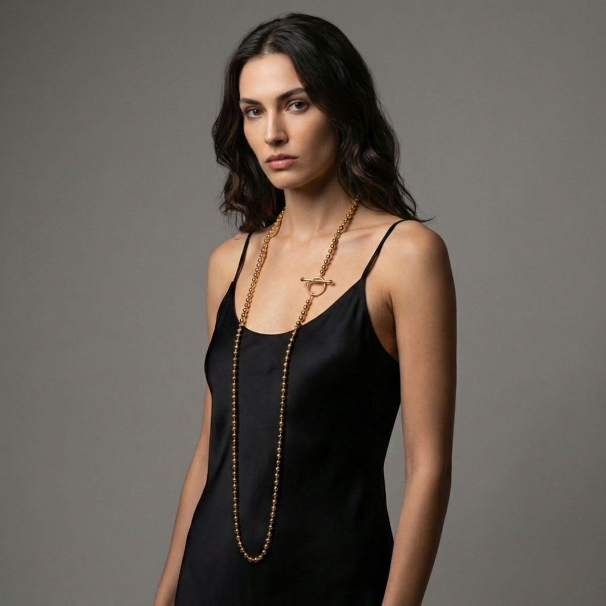 A woman with wavy dark hair wears a sleeveless black dress and the Gold Constellation Orbita Necklace by JAREDJAMIN Jewelry Online, its 18K Gold T-bar clasp gleaming with never-tarnish E-coating as she stands against a plain grey background.
