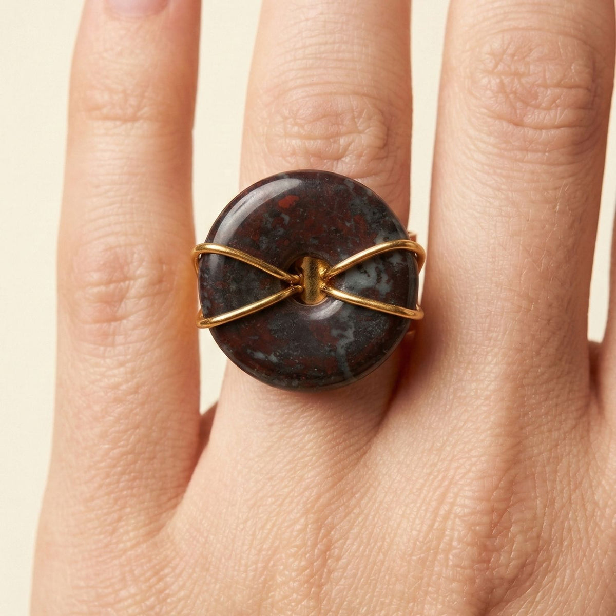 A hand with light skin wears the Gold Cornelia Ring by JAREDJAMIN Jewelry Online, featuring an adjustable 18K gold-plated band with a round, dark Jasper stone showing red and gray patterns. The background is beige.