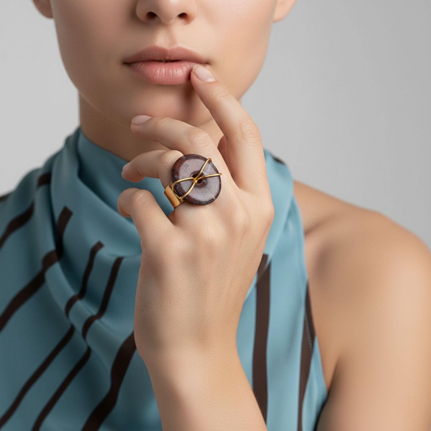 The Gold Cornelia Ring by JAREDJAMIN Jewelry Online features a glossy, round dark jasper bead wrapped in an 18K gold plated wire "X" pattern, creating a minimalistic adjustable statement piece against a plain white background.