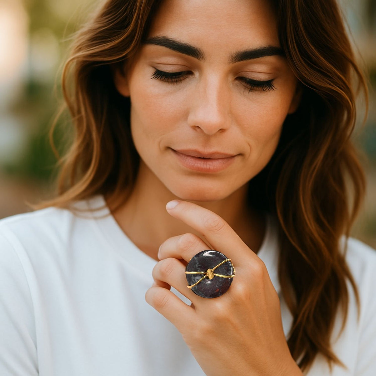 A woman with wavy brown hair and natural makeup wears a white shirt, gently touching her face while showcasing the Gold Cornelia Ring by JAREDJAMIN Jewelry Online—an adjustable statement piece with a large round purple stone on an 18K gold-plated band.