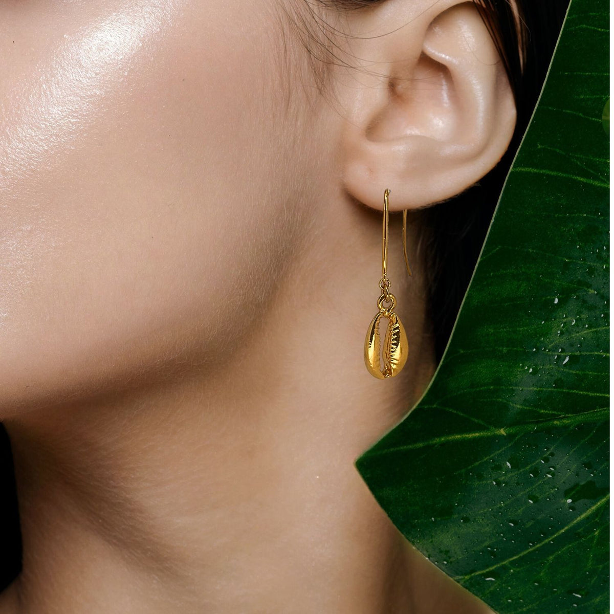 A close-up shows a woman wearing JAREDJAMIN Jewelry Online’s Gold Cowrie French Hook Earrings, her smooth skin glowing under soft light, with a large green leaf dotted with droplets partially covering the image.