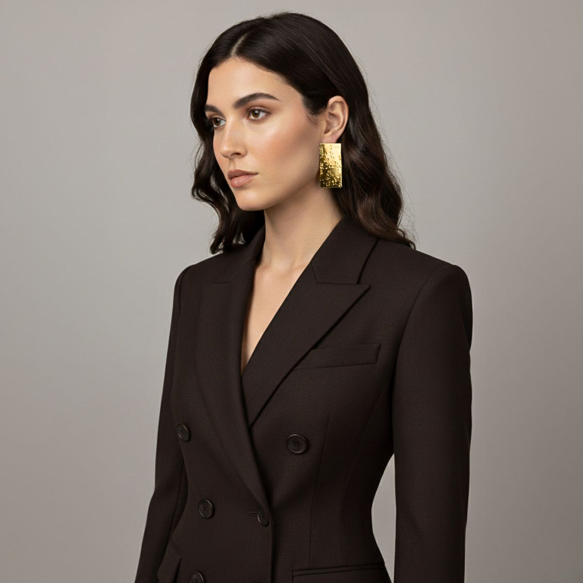 A womanとlong, wavy dark hair wears a dark double-breasted blazerと JAREDJAMIN ゴールド Cuszco Post イヤリング. She poses against a neutral gray background, gazing slightly left 付きの composed expression.