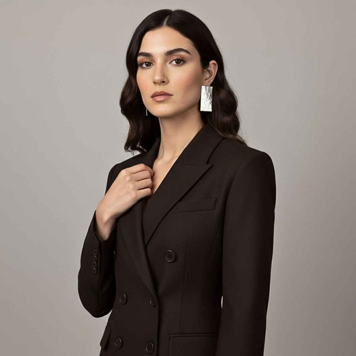 A woman with long, wavy dark hair wears a black double-breasted blazer and JAREDJAMIN Gold Cuszco Post Earrings. Against a neutral background, she gazes confidently at the camera, one hand touching her lapel.