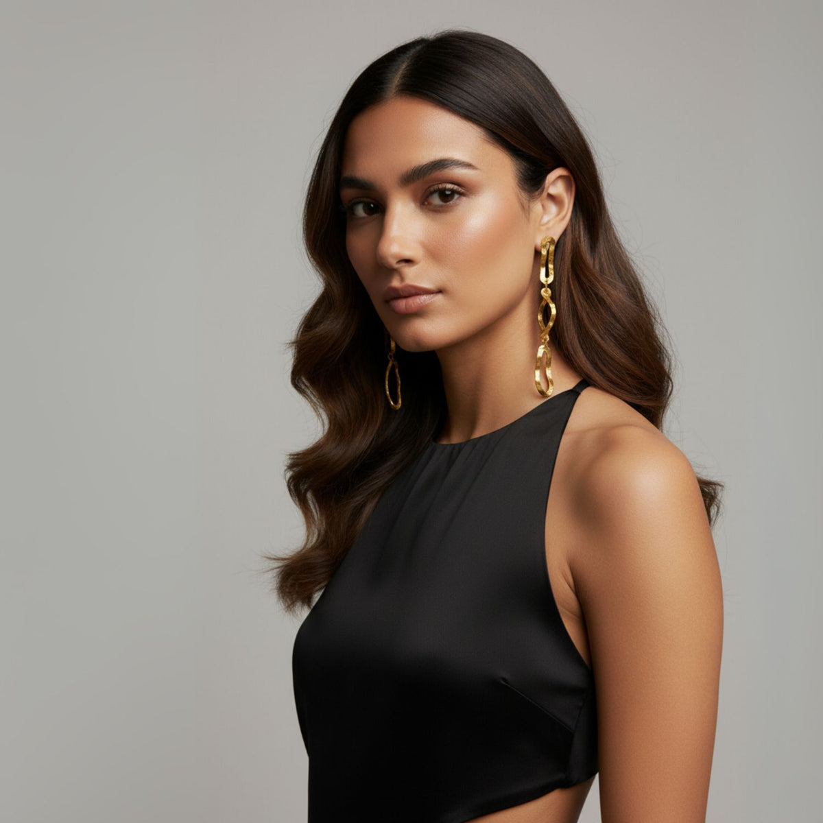 Un woman con wavy dark hair wears a sleeveless black dress y JAREDJAMIN Joyería Online's oro Darla Dangle pendientes. Against a plain background, she gazes confidently at the camera, her natural makeup highlighting her smooth complexion.