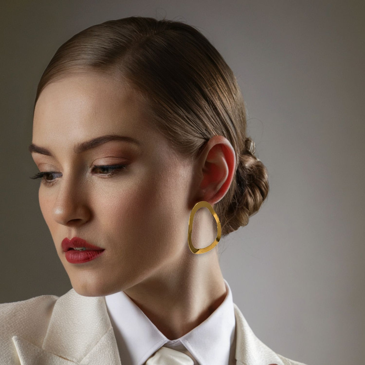 Un woman con fair skin y brown hair en un low bun gazes left, wearing a white blazer, audaz red lipstick, subtle eye makeup, y JAREDJAMIN Joyería Online's 18K oro Plated oro Duet Post pendientes. Los softly lit background is neutral.