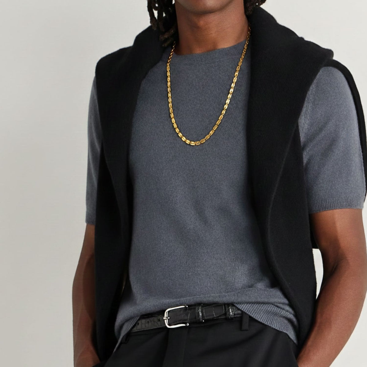 Un person con medium skin tone wears the JAREDJAMIN Joyería Online oro Euclid Rectangular Link cadena collar, a grey short-sleeve sweater, a black sweater over their shoulders, dark braided hair, belted black trousers, y their face partially out of frame.