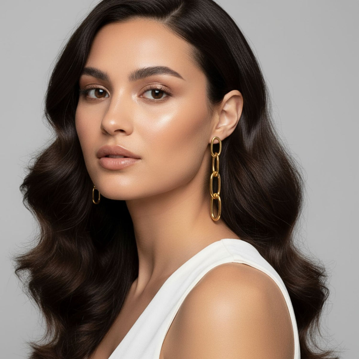 Un woman avec long, wavy dark hair wears a sleeveless white top et or Everly pendante chaîne Post boucles d'oreilles par JAREDJAMIN Bijoux en Ligne, posed against a plain light gray background avec smooth, glowing skin et a neutral expression.
