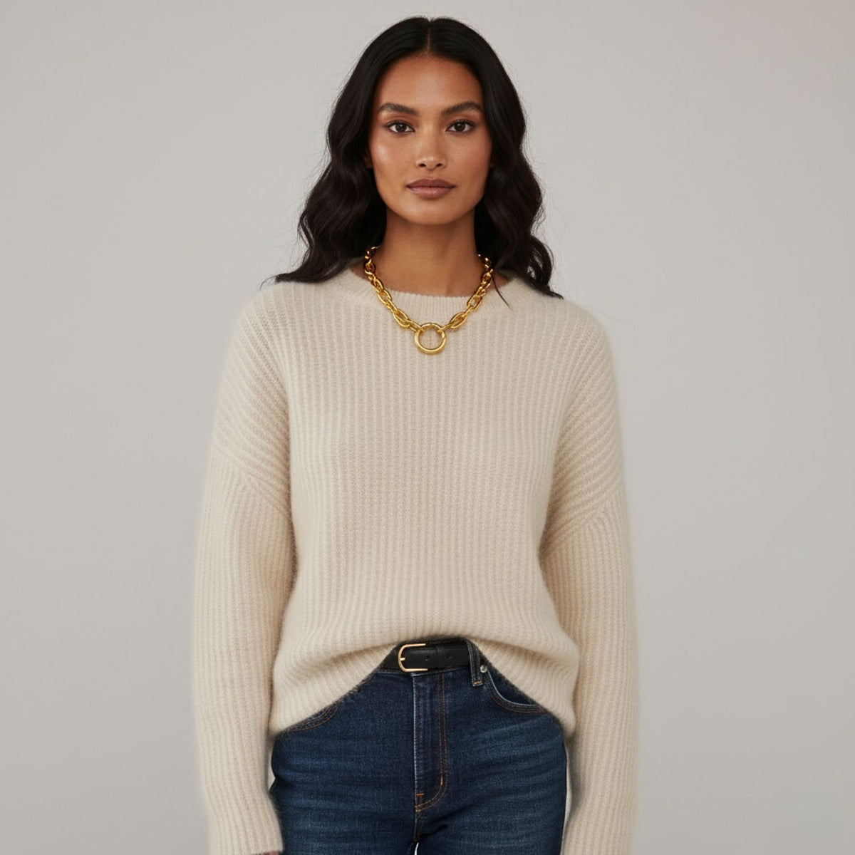 Un woman con long, wavy dark hair wears a cream ribbed sweater, blue jeans con a black belt, y the oro Everly Every-Other collar de JAREDJAMIN Joyería Online. She stands against a light gray background, looking confidently at the camera.