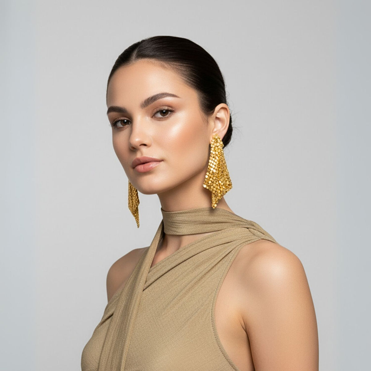 A woman with slicked-back dark hair models JAREDJAMIN Jewelry Online’s Fazzoletto Chainmail Post Earrings, styled with a beige sleeveless crisscross halter top and natural makeup, posing confidently against a plain light gray background.