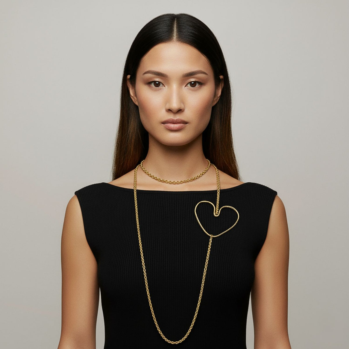 A woman with straight dark hair wears a sleeveless black dress and two gold necklaces: one short, the other a JAREDJAMIN Jewelry Online Gold Fluttering Heart Chain Necklace with a large heart pendant resting on her chest.