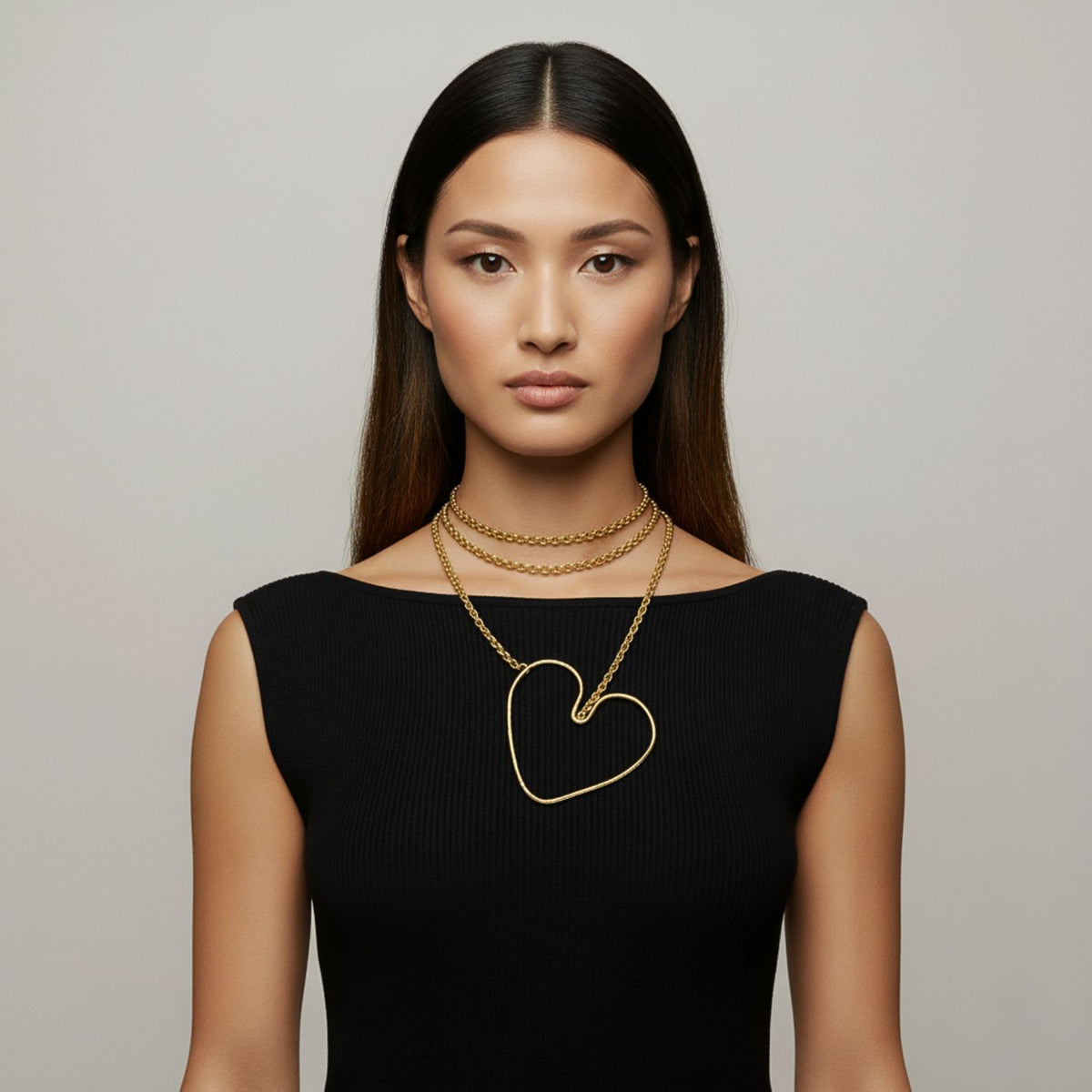 A woman with straight, dark hair wears a sleeveless black top and two gold necklaces: a choker and the JAREDJAMIN Jewelry Online Gold Fluttering Heart Chain Necklace, gazing forward with a neutral expression against a plain light background.