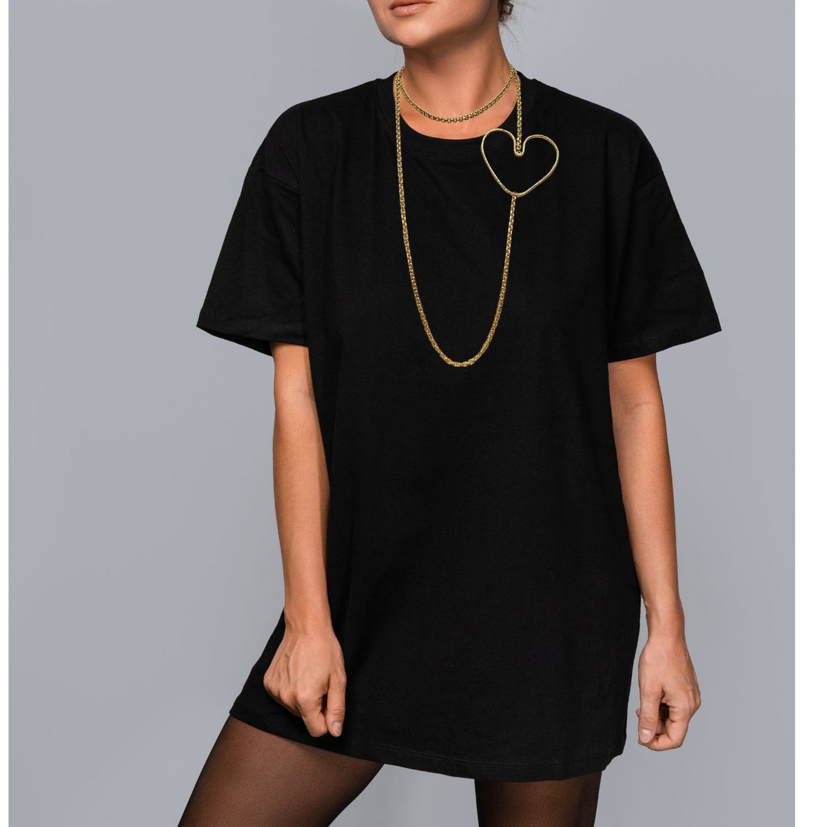 A person in a loose black T-shirt and black tights stands against a gray background, wearing the JAREDJAMIN Jewelry Online Gold Fluttering Heart Chain Necklace. The lobster clasp is hidden, and their face is partially out of frame.