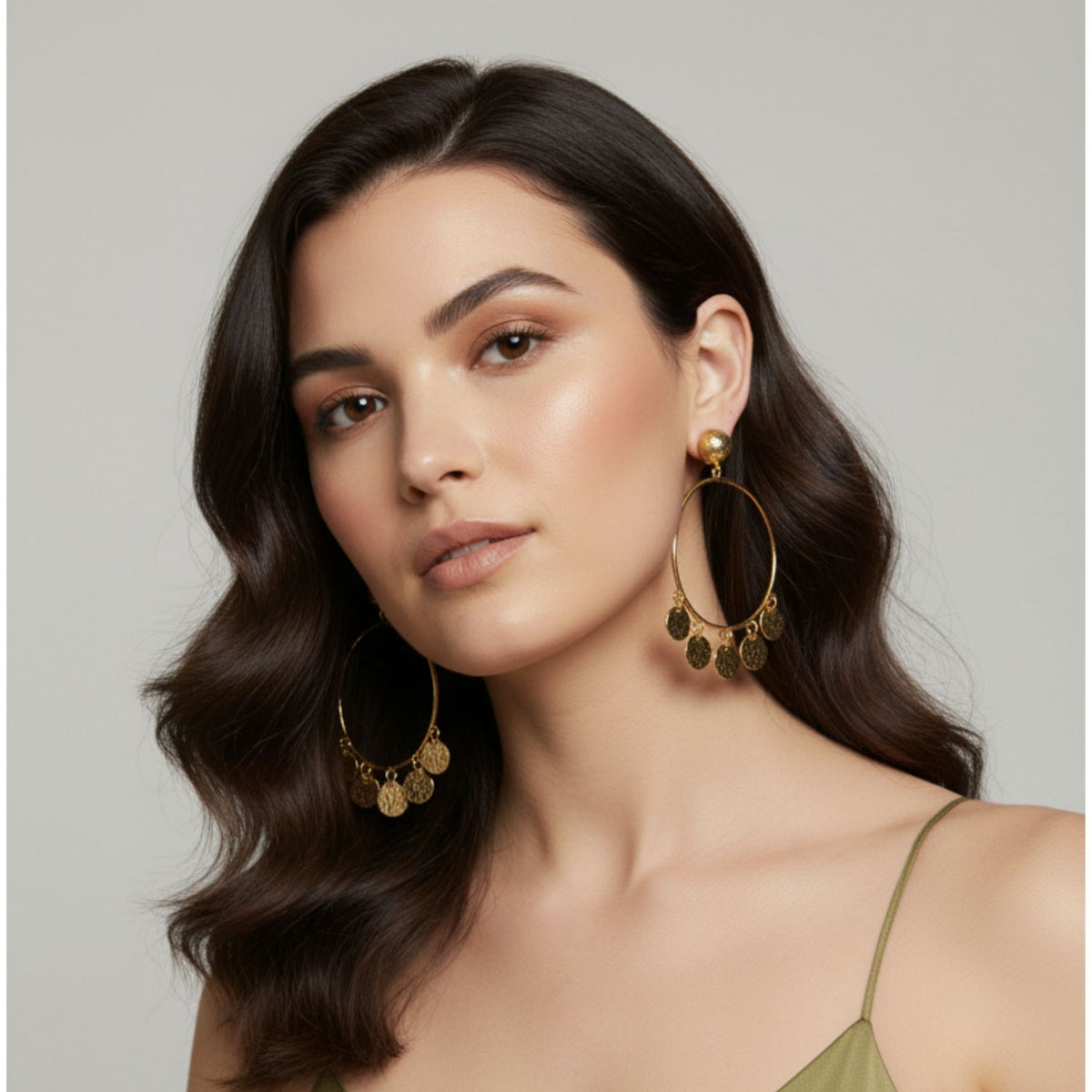Un woman con wavy dark brown hair wears JAREDJAMIN Joyería Online's oro Fortuna Chandelier pendientes y a light olive green spaghetti strap top. Her makeup is natural, con glowing skin y softly defined brows, as she looks calmly at the camera.