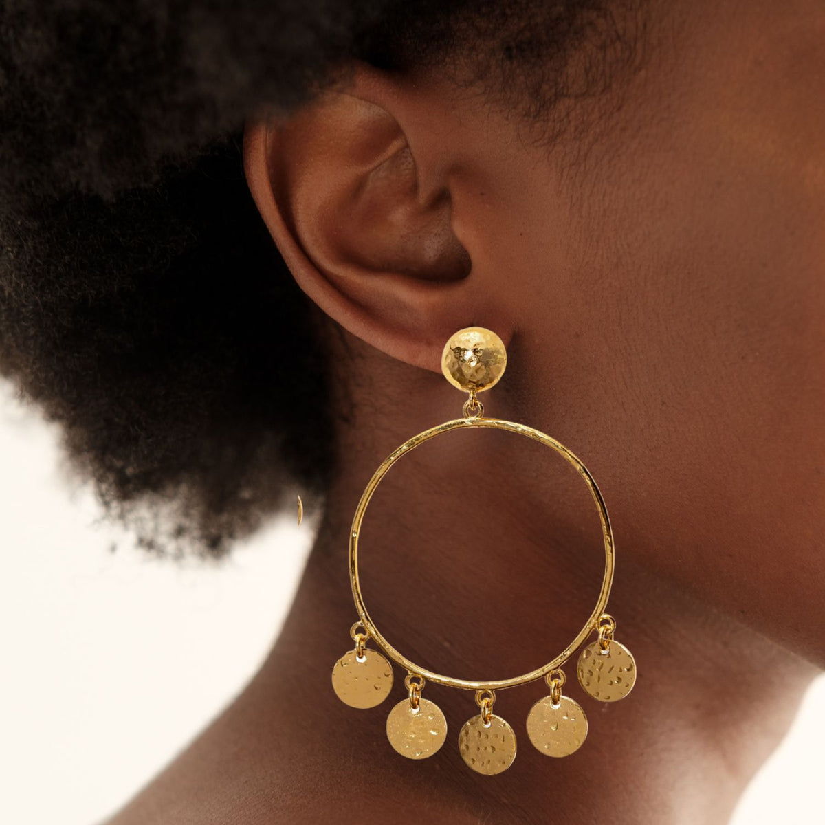 A close-up of a person wearing JAREDJAMIN Jewelry&#39;s Gold Fortuna Chandelier Earrings, elegantly plated in 18k gold. The large hoop showcases five small, textured discs with a shimmering e-coated finish and is attached by a gold stud, beautifully complementing the person&#39;s short, curly hair.