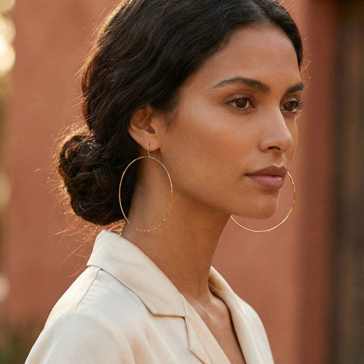 A woman with medium skin and dark wavy hair in a low bun wears JAREDJAMIN Jewelry Online’s Gold Halo Textured Hoop Earrings and a cream blouse, standing outdoors against a blurred, warm-toned background.