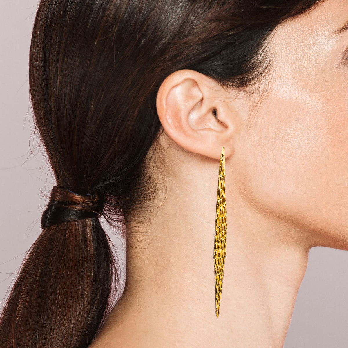 A woman with straight, dark brown hair in a low ponytail is shown in profile, wearing JAREDJAMIN Jewelry Online&#39;s Jaguaretê Earrings with 18K Gold and Silver Plating. The plain background highlights her smooth skin and the earrings&#39; striking design.